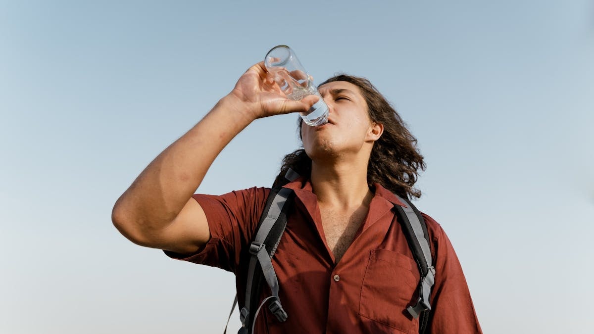 A young man in a red shirt drinks water from a bottle under a clear sky, highlighting hydration and outdoor adventure.